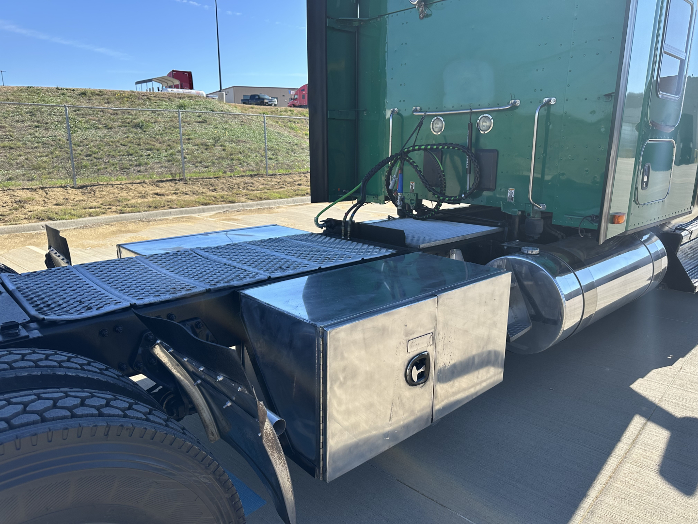 Kenworth T680 tractor with stainless steel toolbox mounted behind the cab, fuel lines and exhaust visible; side view showing green sleeper cab and rear wheel area.