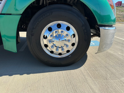 Front wheel of a green 2023 Kenworth T680 with a shiny chrome hub and Michelin tire; partial front fender and bumper visible.