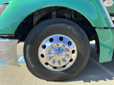 Front wheel and front fender of a green 2023 Kenworth T680 undercarriage. Wheel shows a polished chrome hubcap with lug nuts; tire sidewall reads KENWORTH and other markings.
