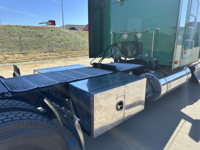 Kenworth T680 tractor with stainless steel toolbox mounted behind the cab, fuel lines and exhaust visible; side view showing green sleeper cab and rear wheel area.
