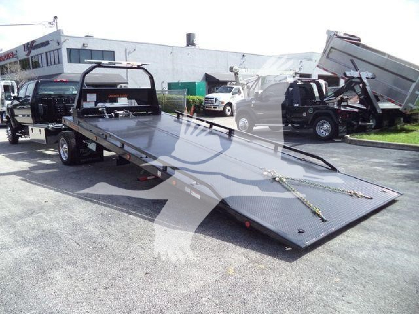 Flatbed tow truck with a lowered ramp, International CV series rollback truck in background, lot with other trucks.