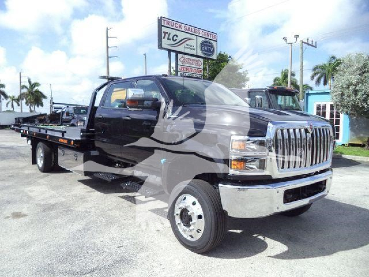 Truck showing a flatbed rollback with a chrome grille and dual exhaust; TLC Truck Sales Center sign in background.