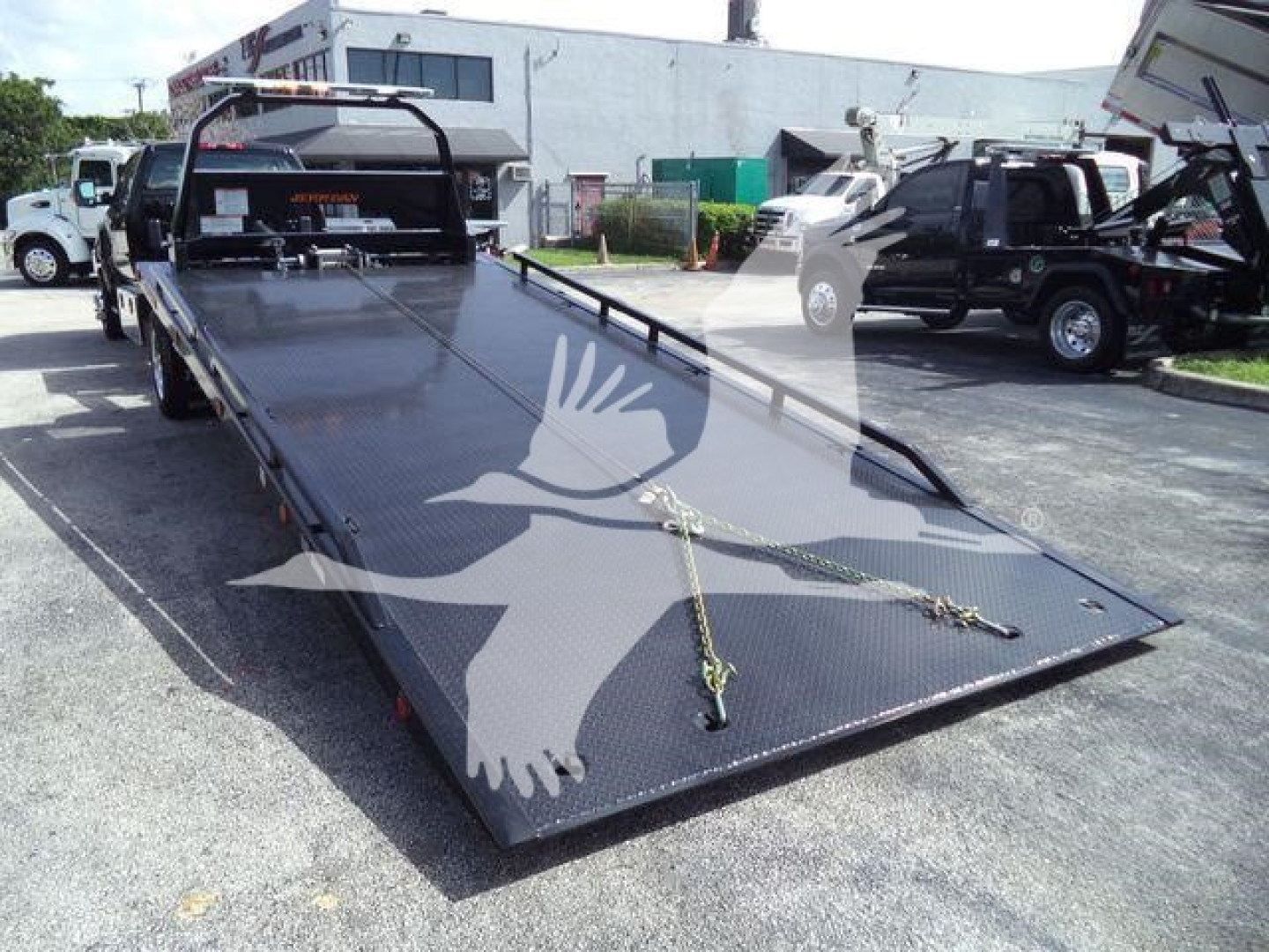 Tow truck rollback flatbed with loading ramps and winch lines, viewed from front-left corner, in a dealership lot.