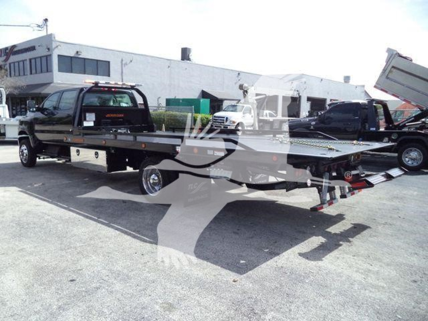 Flatbed rollback tow truck from side, black cab, long bed with ramps and wheel lift, outdoor lot with other trucks in background