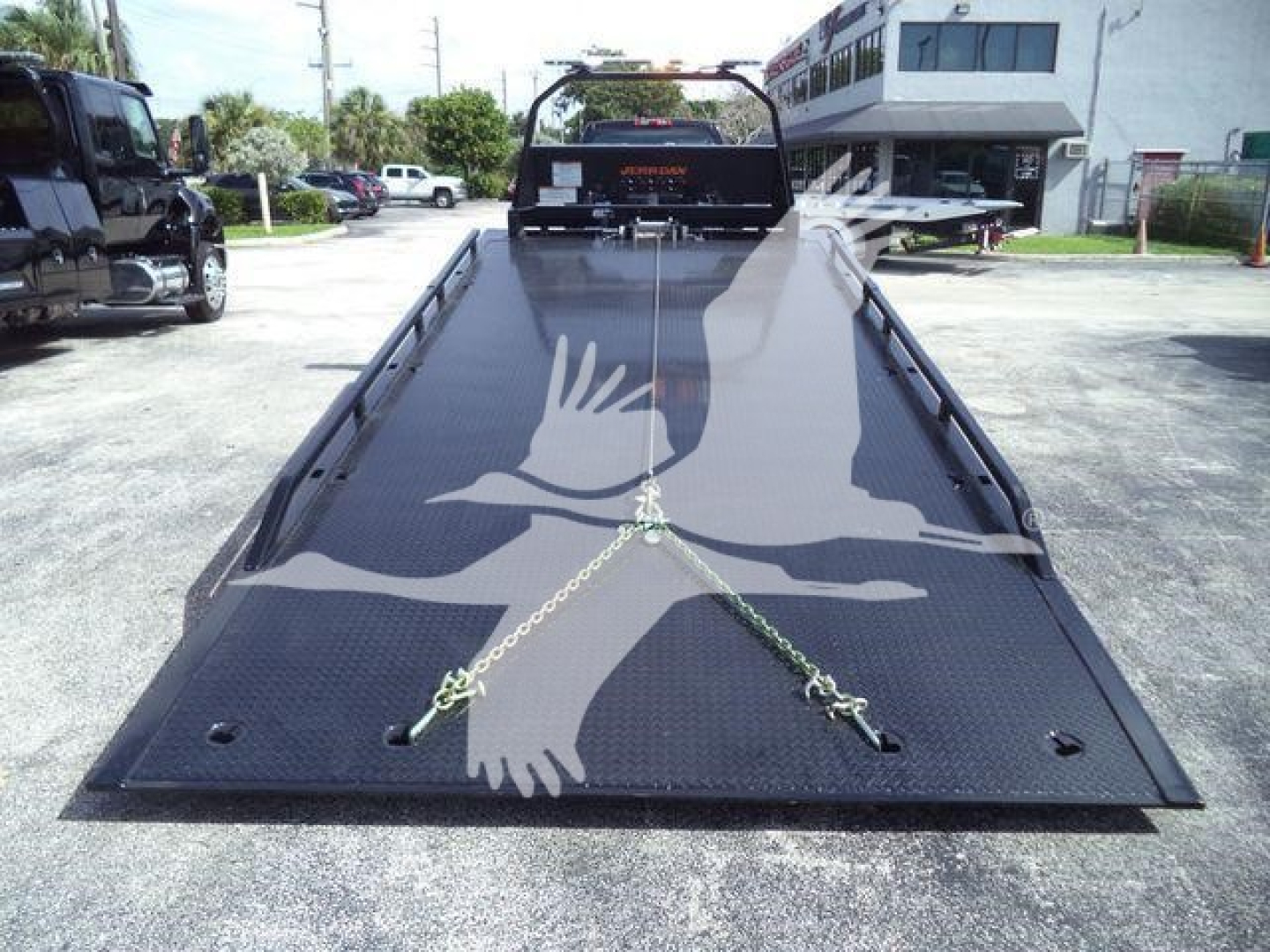 Wide view of a black flatbed tow truck bed (rollback) with chains secured in front; background shows dealership building and other trucks.