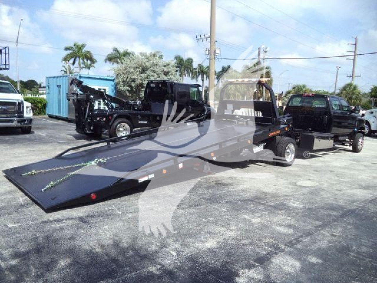 Truck with rollback flatbed in serviceable condition, International CVW or FV-style chassis, blue sky, palm trees, multiple vehicles in background. Text not visible.