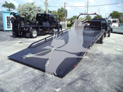 Flatbed tow truck trailer with hydraulic tilt deck and wheel straps in a dealership lot.