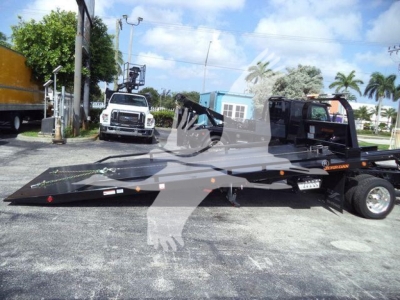 Tow truck rollback flatbed with black metal deck and wheel chocks, International CV rollover truck in background, vehicle dealership lot.