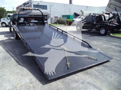 Tow truck rollback flatbed with loading ramps and winch lines, viewed from front-left corner, in a dealership lot.