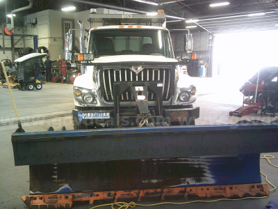 Front view of a white 2010 International WorkStar 7400 dump truck in a shop with a blue snow plow blade attachment in the foreground.