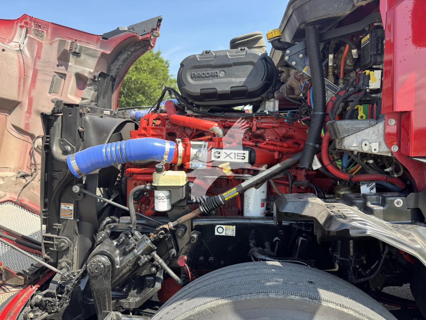 Kenworth T680 engine bay showing bright red diesel engine with blue radiator hose, air intake, and various hoses and wiring; visible components include air filter housing and coolant reservoir.