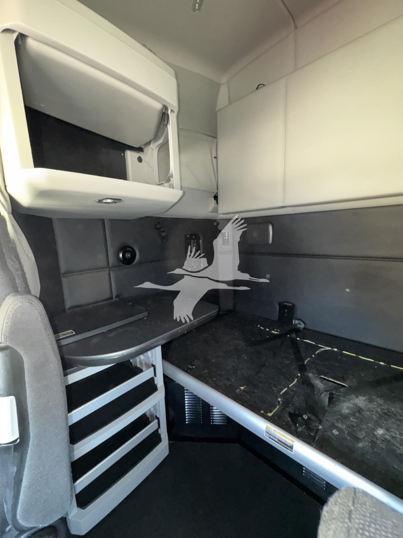 Kenworth T680 interior sleeper cab area with upper bunk storage and seating/display area; visible are a small cabinet, overhead shelf, and partition wall.