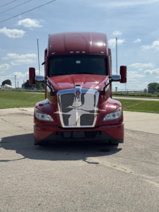 Kenworth T680 front view showing prominent chrome grille with large hood, maroon exterior, dual mirrors, daytime running lights, and highway background.