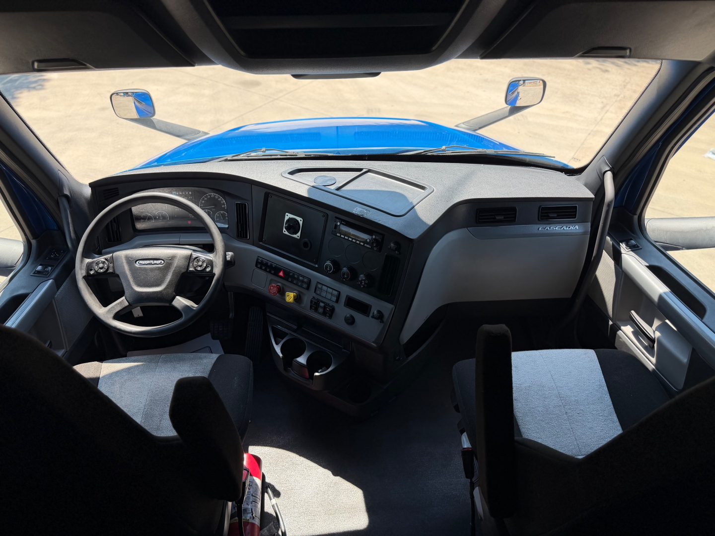 Cab interior of a Freightliner truck showing steering wheel, dashboard with analog gauges, center console, and seating; blue exterior seen through windshield.