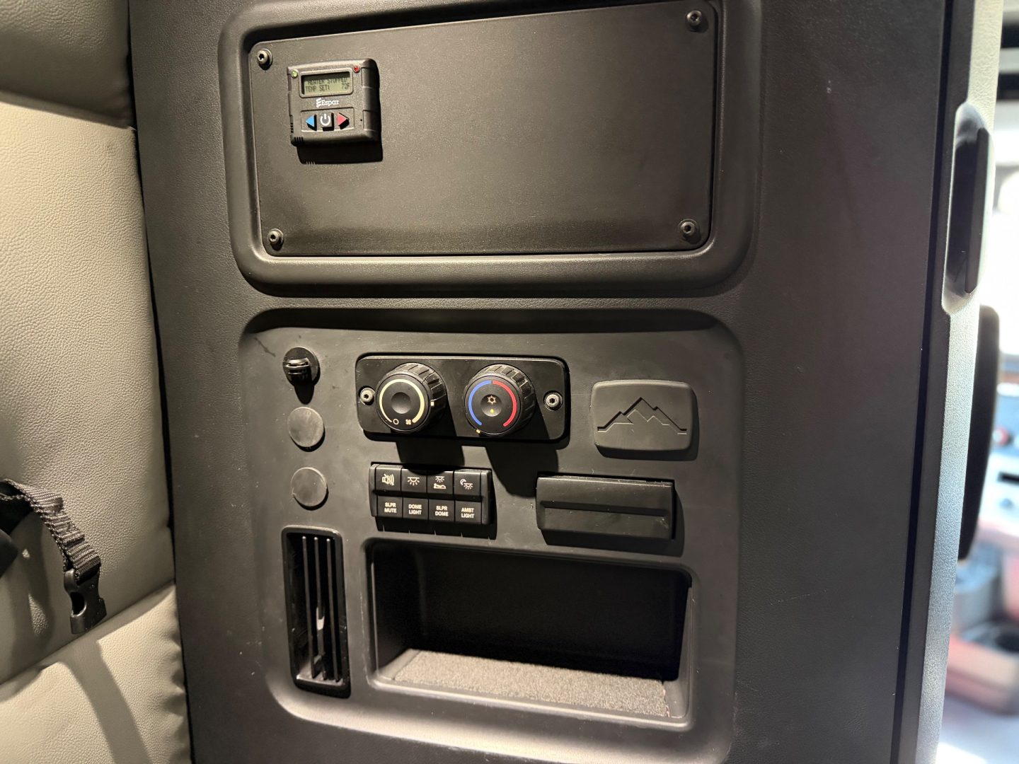 Dashboard area of a Freightliner truck cab with knobs, vents, and a small cubby; visible are circular climate control dials, a USB-like port panel, and a rectangular storage slot.