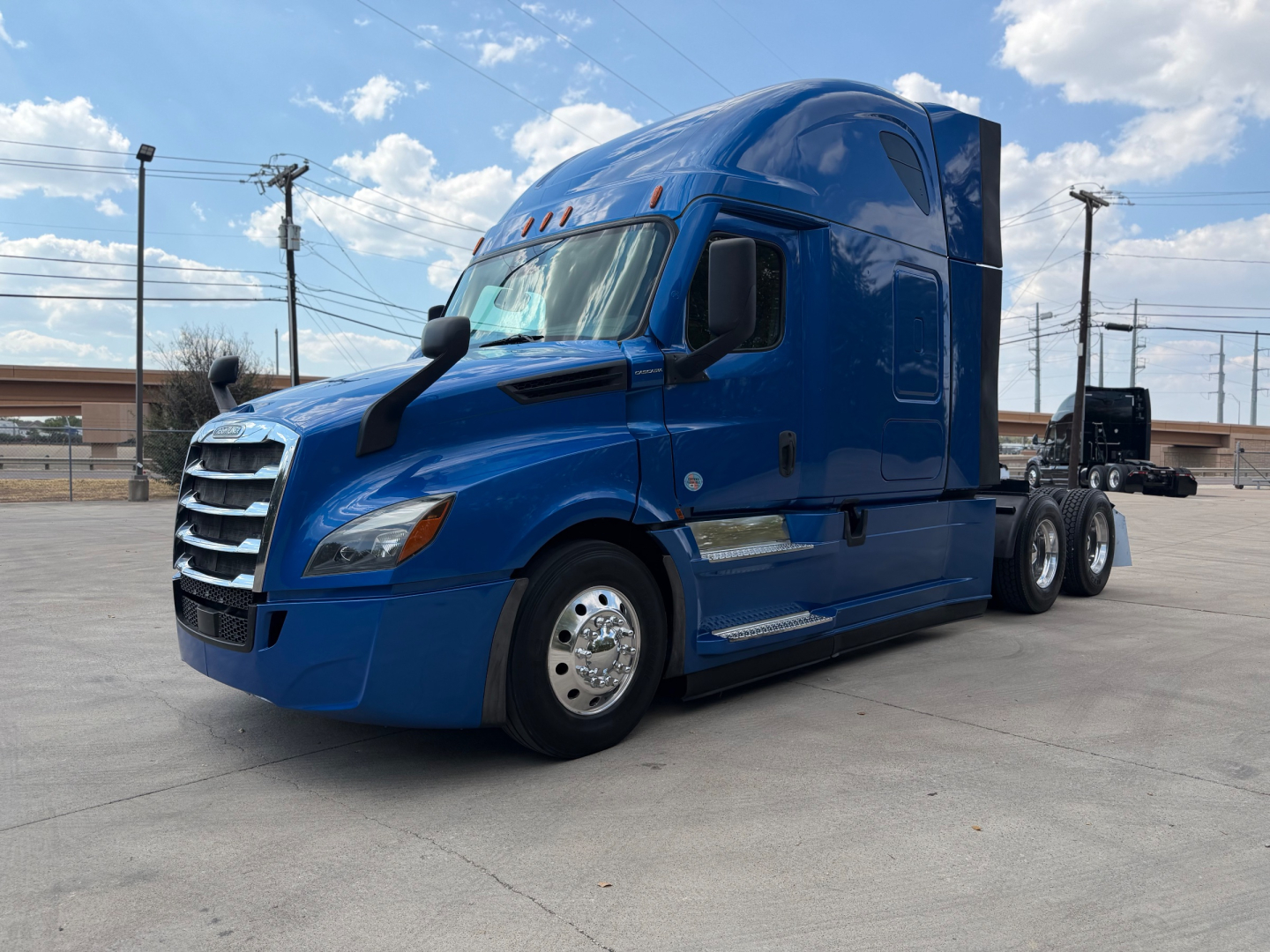 Freightliner tractor cab in blue, side view showing sleeper configuration and dual rear axles on a concrete lot.