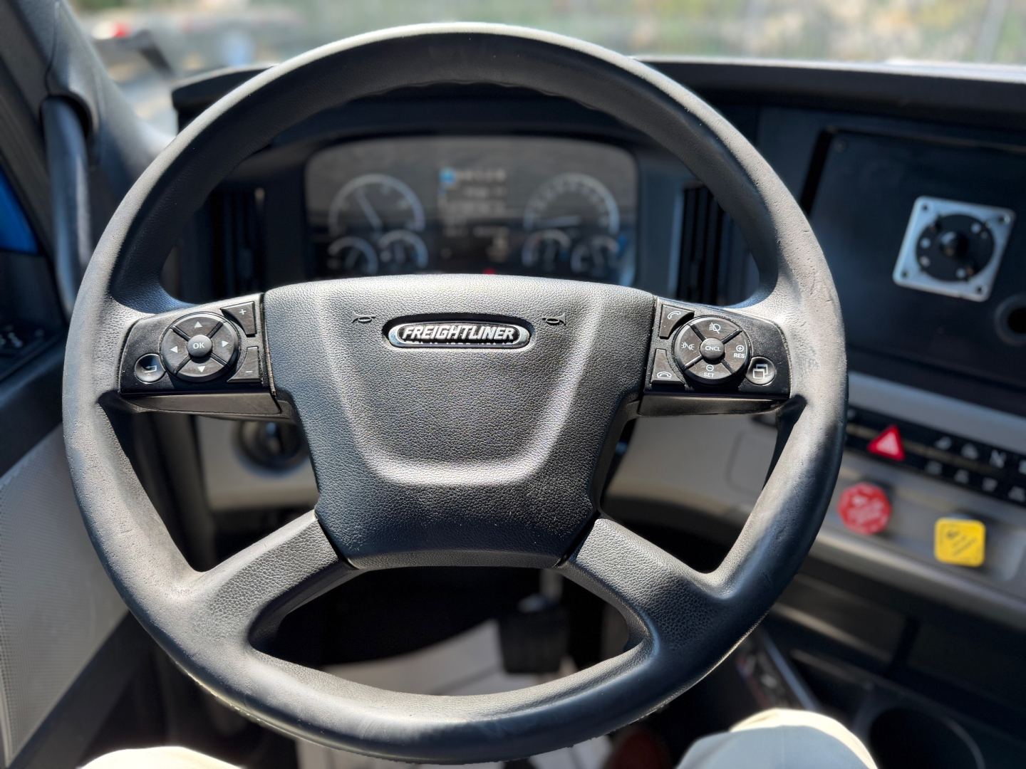 Freightliner steering wheel in cab with dashboard gauges visible; circular controls on wheel, center badge reads Freightliner.