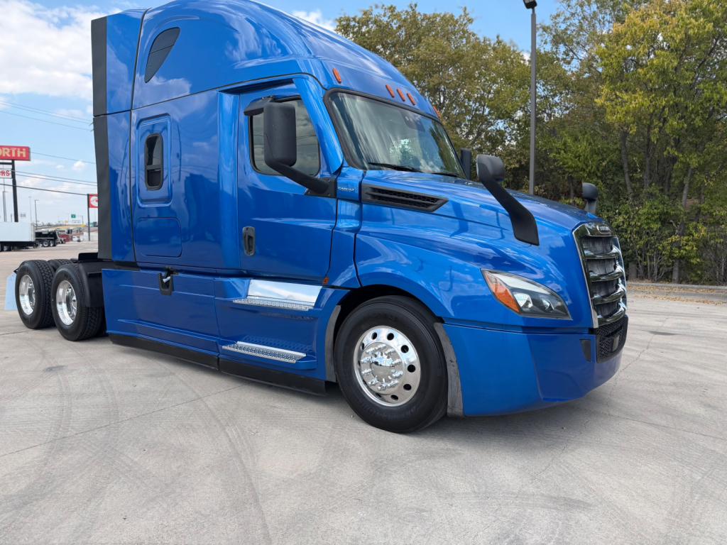 Truck cab in blue showing Freightliner PT126064ST sleeper tractor with aerodynamic design, large side mirrors, chrome fuel tanks, and dual rear axles; front grill and headlights visible.