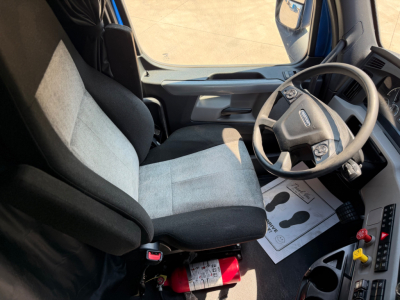 Cab interior of a Freightliner truck showing steering wheel, driver's seat, and dashboard. Visible are red fire extinguisher near the seat, floor mat with printed outlines, and control panel with colored knobs.