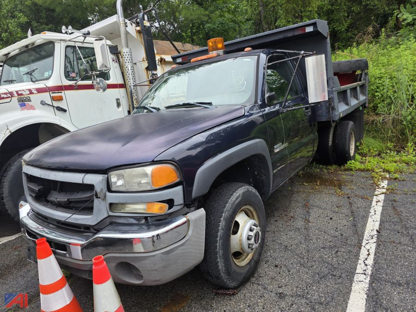 GMC Sierra 3500 dump truck with plow attachment, dark blue cab, front view, single rear axle, utility bed, on a parking lot with traffic cones nearby.