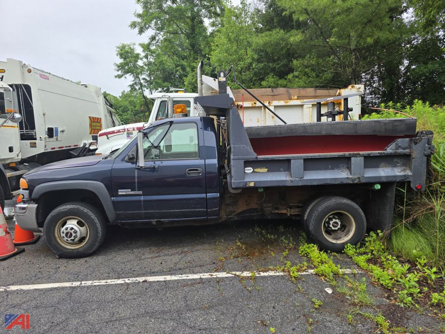 2006 GMC Sierra 3500 dump truck with plow, dark blue cab, front wheel drive, dump bed raised slightly, roadside gravel lot, weeds along edge.