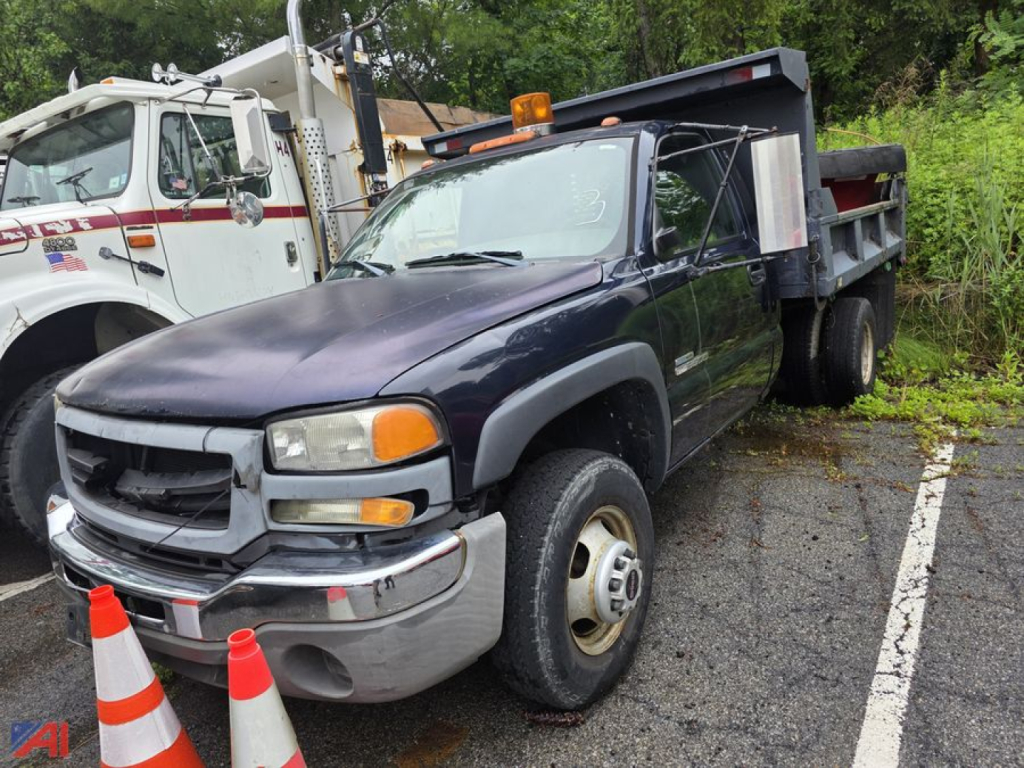 GMC Sierra 3500 dump truck with plow attachment, dark blue cab, front view, single rear axle, utility bed, on a parking lot with traffic cones nearby.