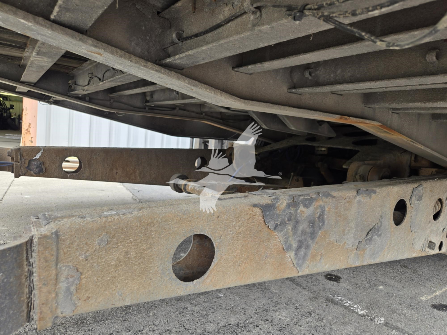 Trailer undercarriage showing rusted steel frame and mounting brackets with a cross member and holes, undercarriage components visible.