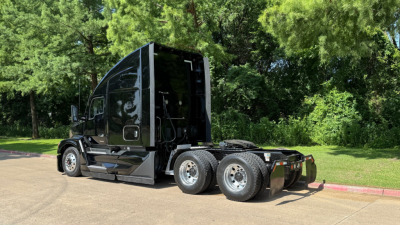 Truck chassis and cab of a 2023 Kenworth T680 conventional sleeper viewed from rear side showing dual rear axles and fuel/wuel lines, chrome wheels, and black paint.