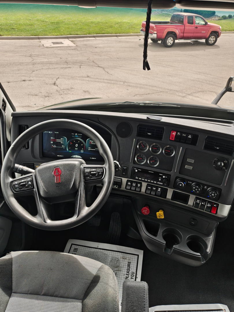 Kenworth T680 cab interior dashboard and steering wheel with controls visible, truck windshield showing outside red pickup and parking lot.