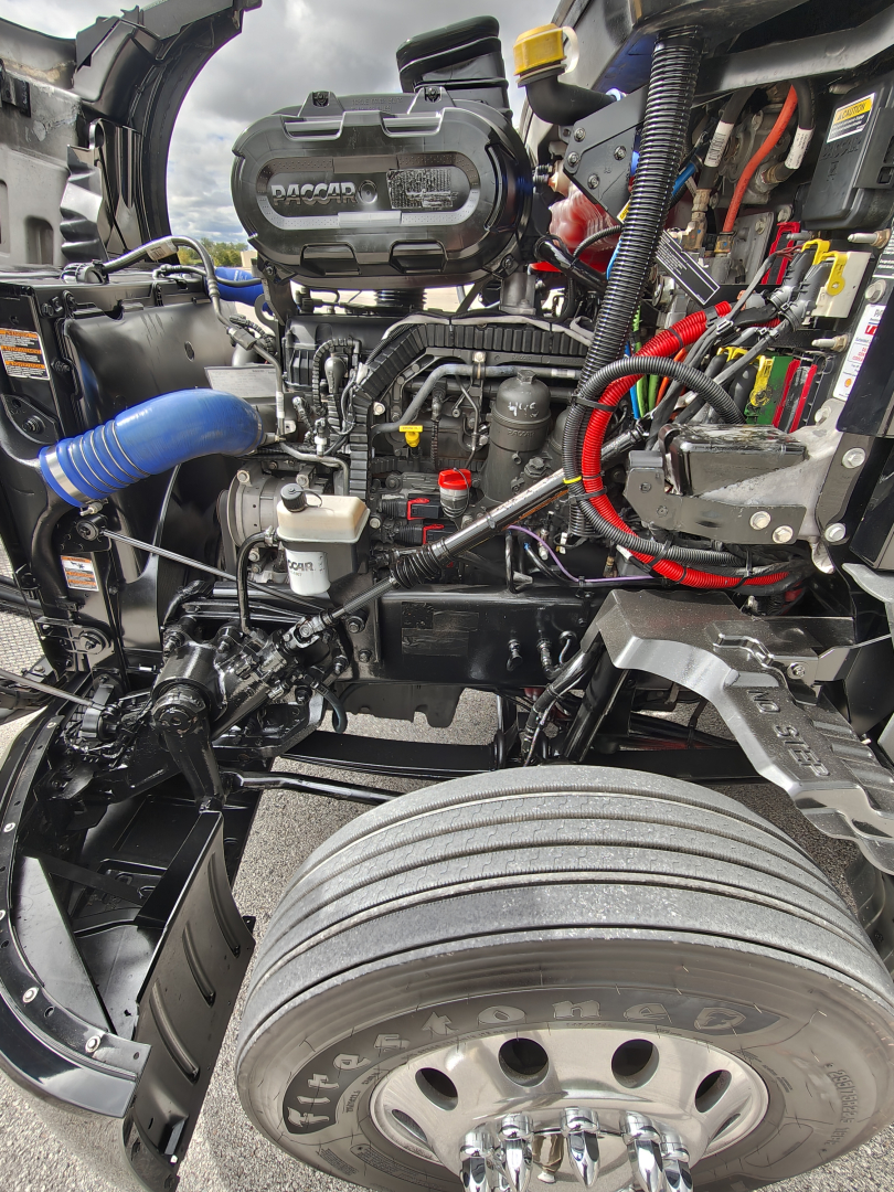 Kenworth T680 engine bay view showing engine components and wiring with a front wheel in frame.