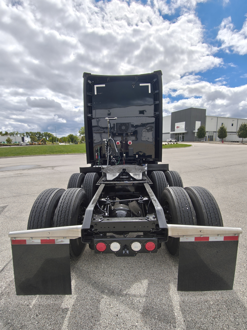 Rear view of a Kenworth truck chassis with fifth-wheel and dual rear tires, black cab section raised, rear lights and mudflaps visible.