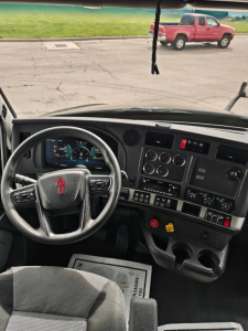 Kenworth T680 cab interior dashboard and steering wheel with controls visible, truck windshield showing outside red pickup and parking lot.
