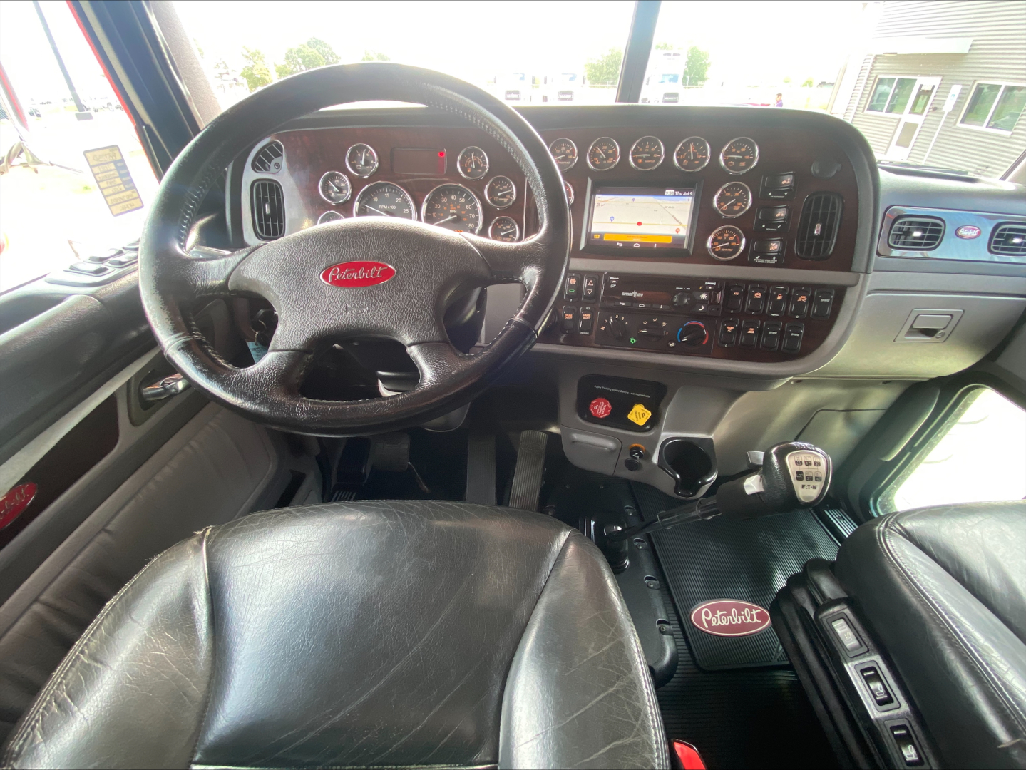 Peterbilt 389 dashboard and steering wheel in-cab view showing instrument cluster, center console with touchscreen, and gear shifter; black leather seats; Peterbilt logo on floor mat and steering wheel.
