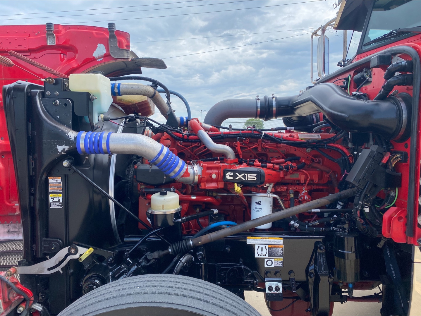 Peterbilt 389 engine bay showing Cummins X15 engine with blue silicone hoses, radiator, coolant reservoir, and various hoses in red truck chassis.