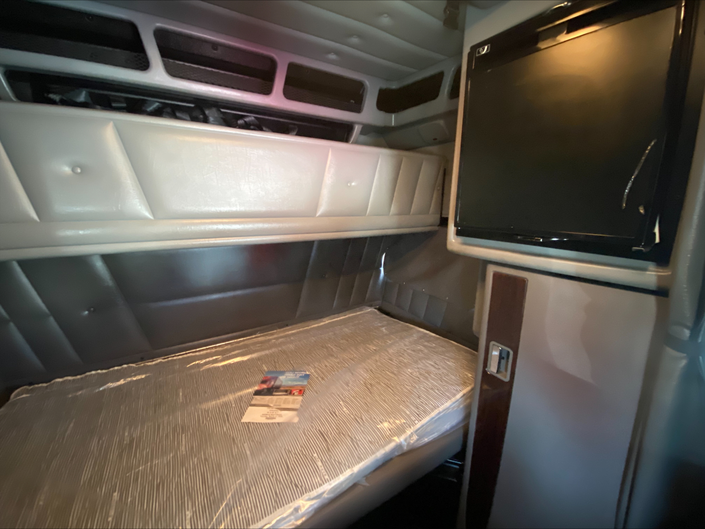 Peterbilt truck cab interior with bunk bed and small fridge, visible vents and overhead storage; no legible text on components.