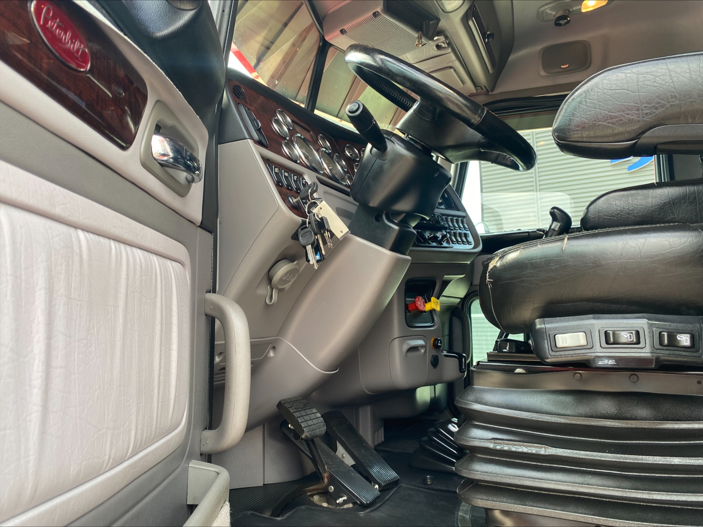 Peterbilt 389 interior cabin view showing steering wheel, dashboard, pedals, and driver's seat with control switches.