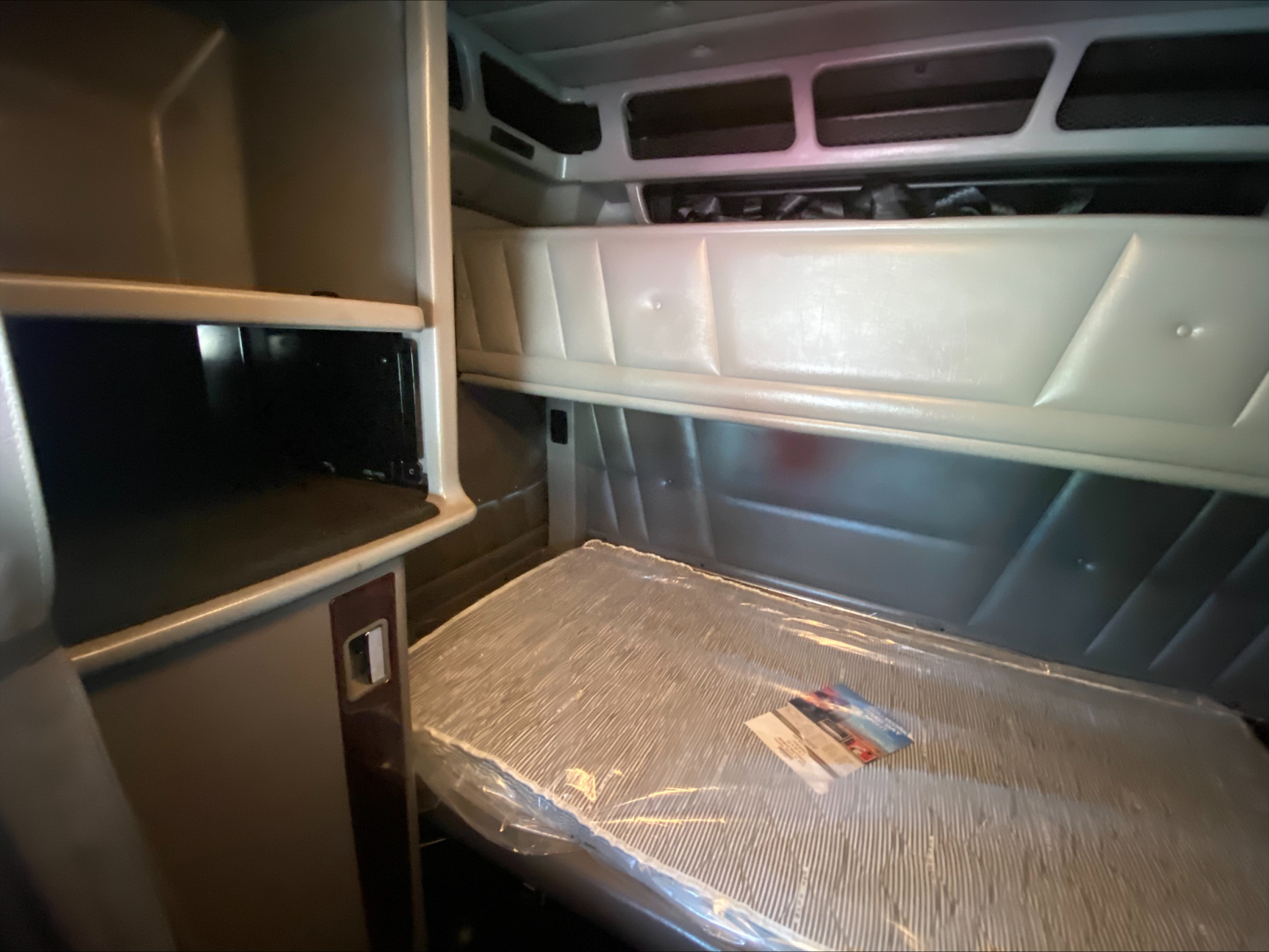 Peterbilt 389 sleeper cab interior shelf and dash area showing storage cabinet and window panel with plastic-wrapped floor mat.