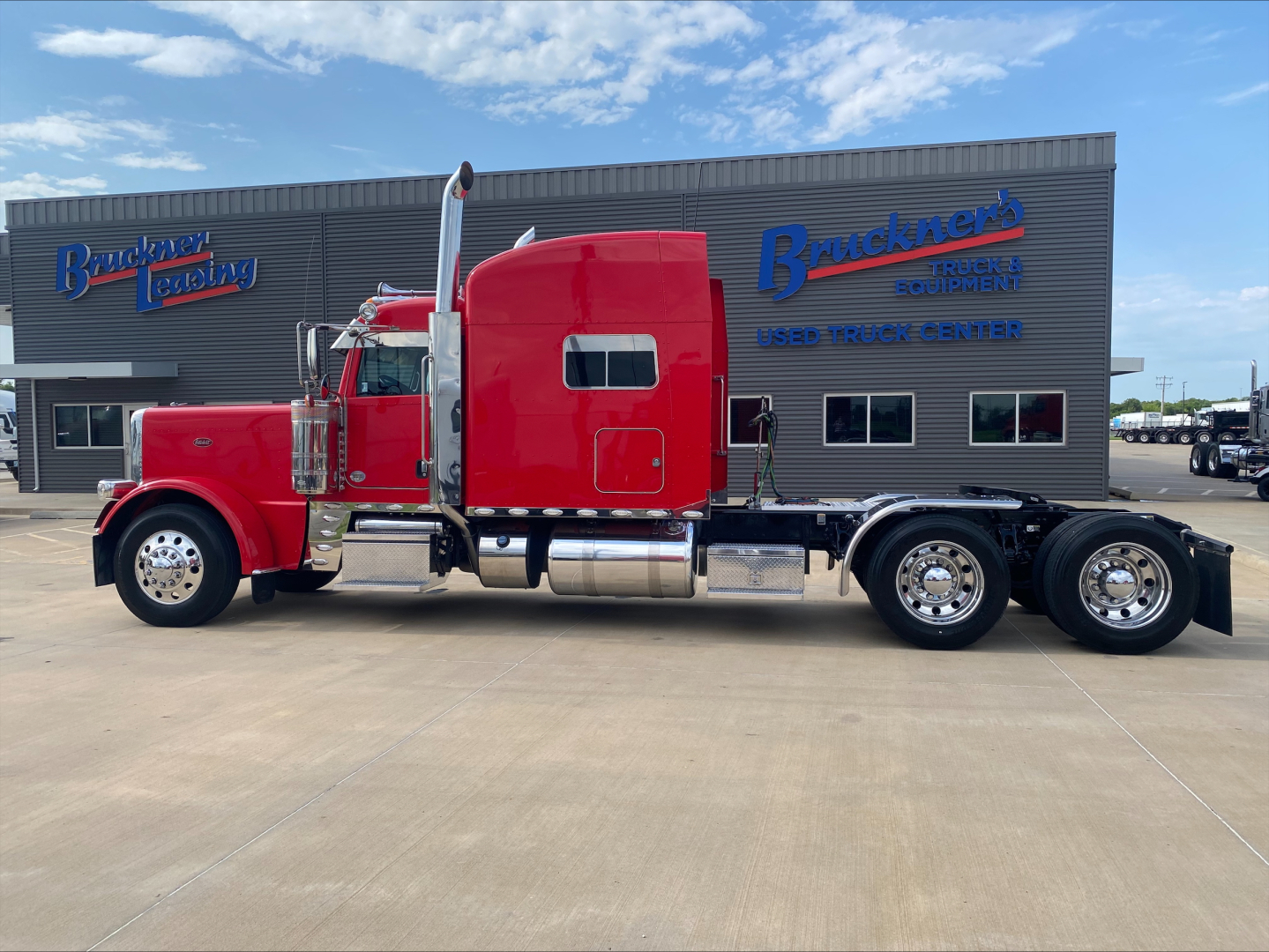 Red Peterbilt 389 conventional sleeper truck side view with chrome fuel tanks and triple rear axles parked in front of a Bracken dealership building.