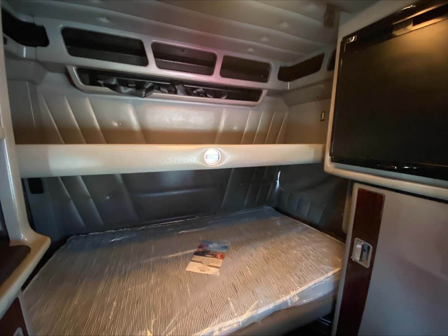Peterbilt 389 sleeper cab interior showing overhead dash area and bed with plastic covering and a small booklet on the mattress.