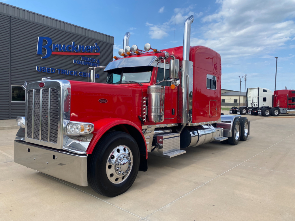 Peterbilt 389 conventional sleeper truck in red with chrome grille and dual exhaust stacks, parked outside dealership