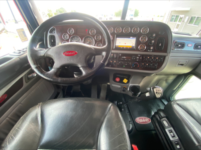 Peterbilt 389 dashboard and steering wheel in-cab view showing instrument cluster, center console with touchscreen, and gear shifter; black leather seats; Peterbilt logo on floor mat and steering wheel.