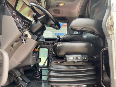 Interior of a Peterbilt 389 cab with black leather driver seat and steering wheel, dashboard cluster, and controls visible; gear shifter and pedals in view.