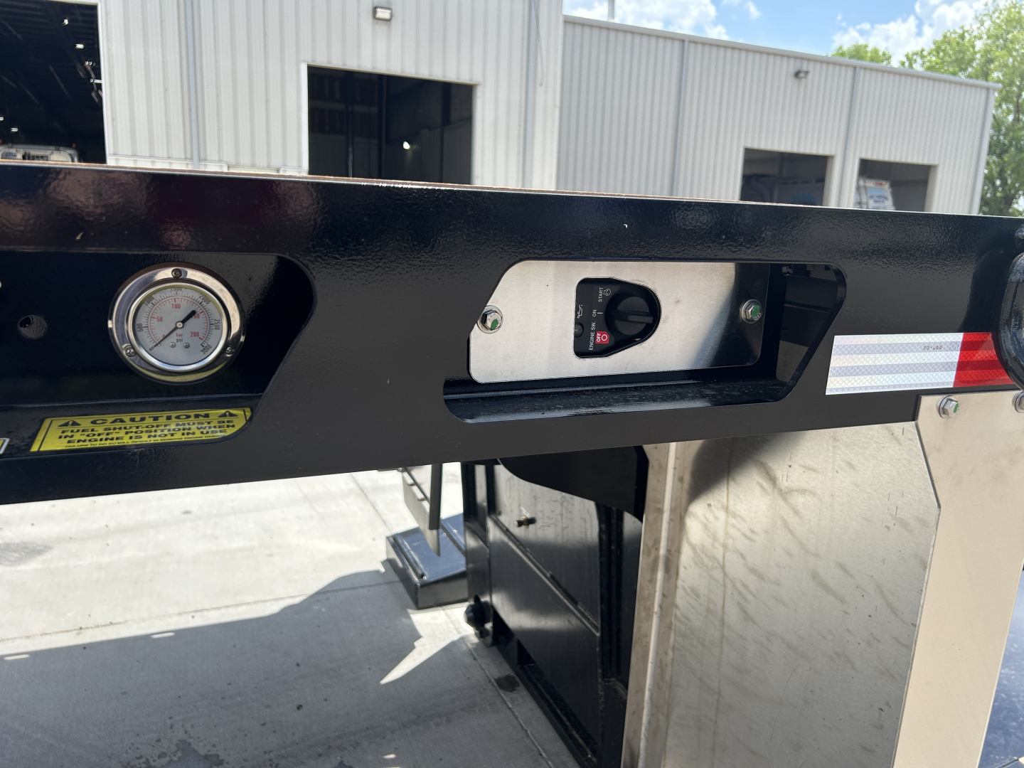 Table/contour of a trailer rear with a gauge, a switch panel, and a safety label. Visible text includes a round gauge, a small rectangular switch area, and a warning label. The frame is black with a white/red reflective strip.