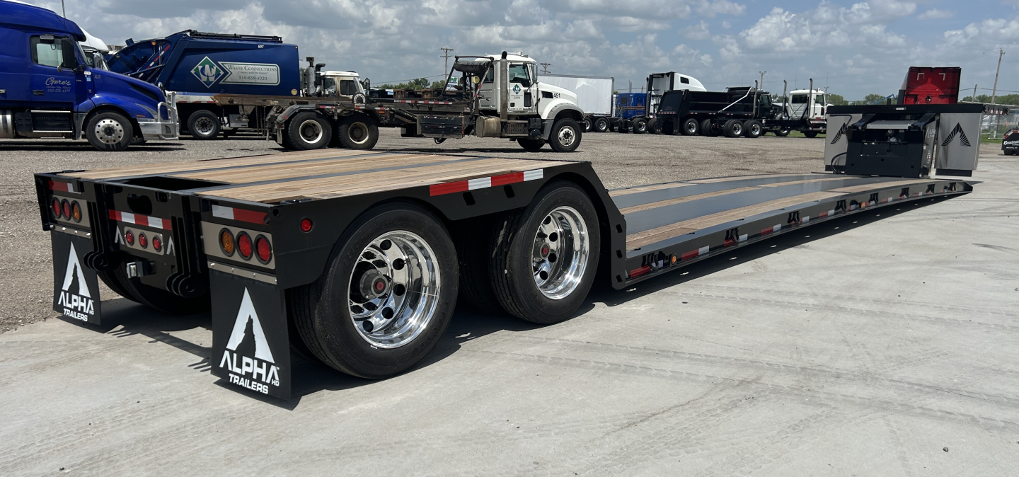 Trailer showing long lowboy platform with dual rear axles and aluminum wheels; Alpha logo mud flaps; various lights and reflectors on the rear; hydraulic detatch and double drop mini deck trailer configuration.
