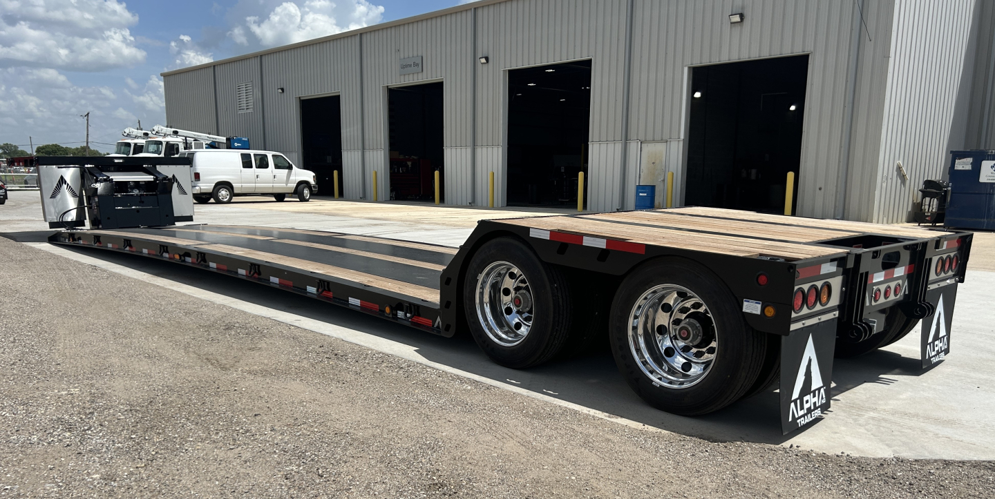 Trailer in a lowboy configuration showing a long flatbed with dual rear axles and slick tires.