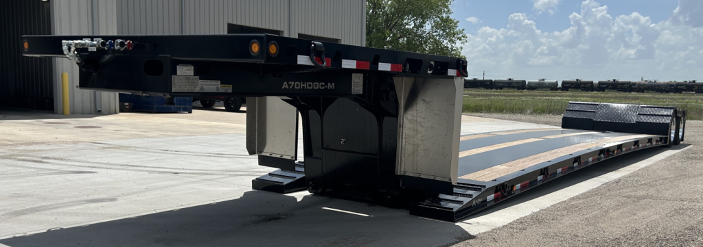 Lowboy trailer in yard showing hydraulic detachable double drop mini deck with long flat bed and gooseneck area. Clear weather, rear view with ramp sections and side reflectors.