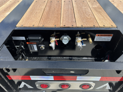 Trailer deck area showing hydraulic detent controls and gauges on a lowboy trailer bed, with wood deck planks and metal frame, warning label visible.