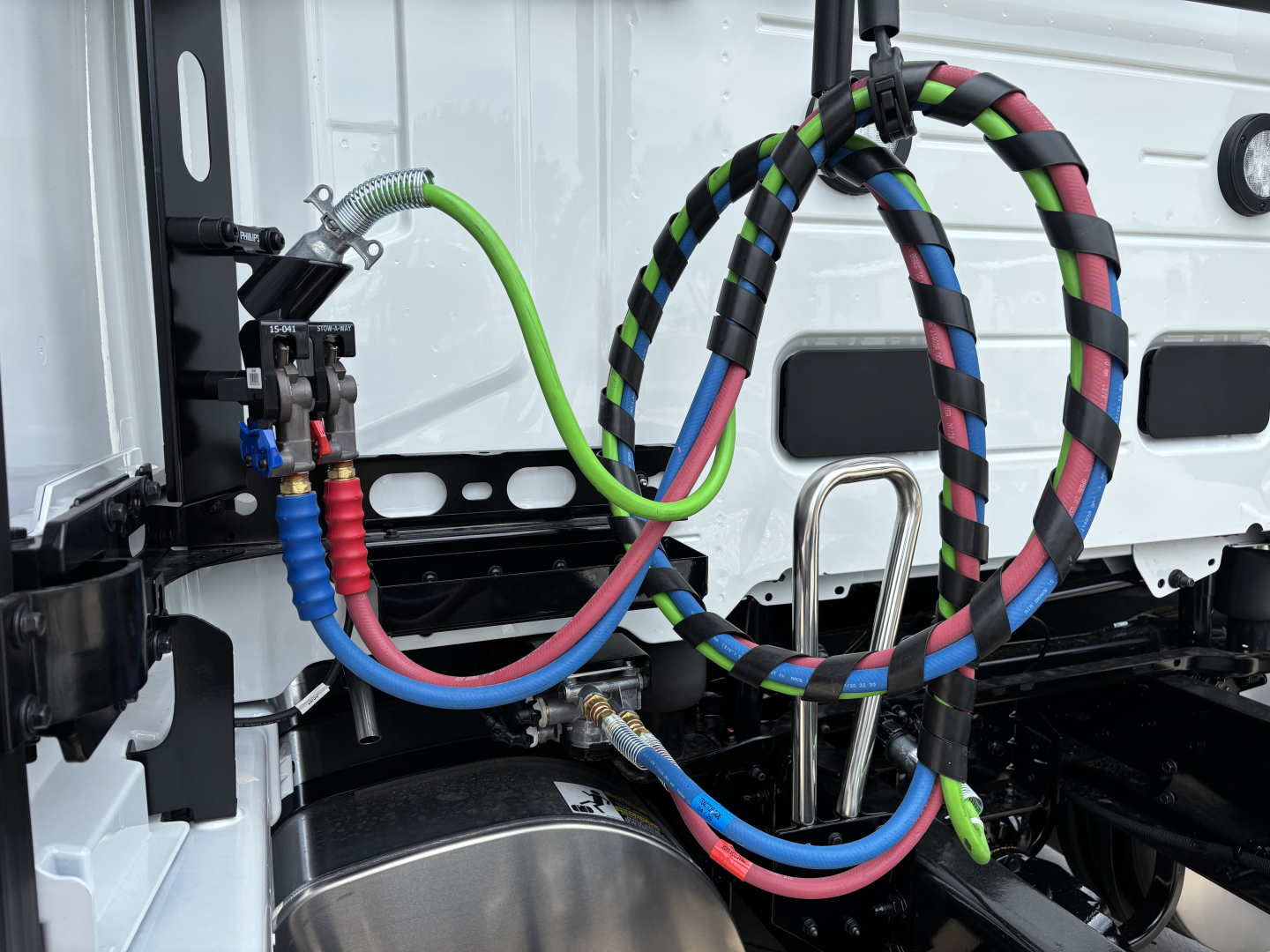 Volvo VNL chassis with bundled hydraulic lines and cables; visible color-coded hoses (green, red, blue, black) connected near the cab mount; metal brackets and exhaust area