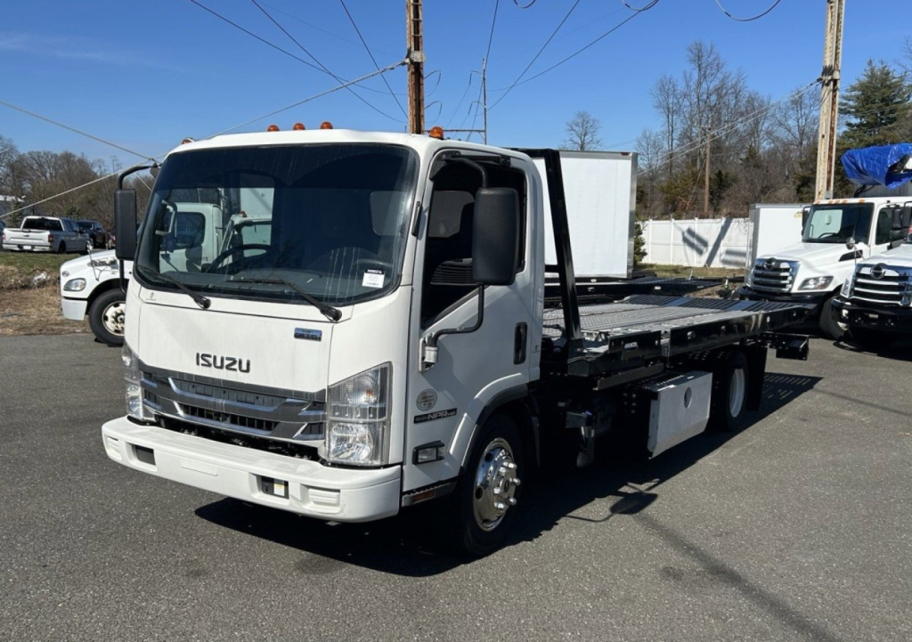 Isuzu NRR box truck with a tilt-bed or rollback tow setup in a dealership lot, white cab, front view, JALE5W167L7306216 visible