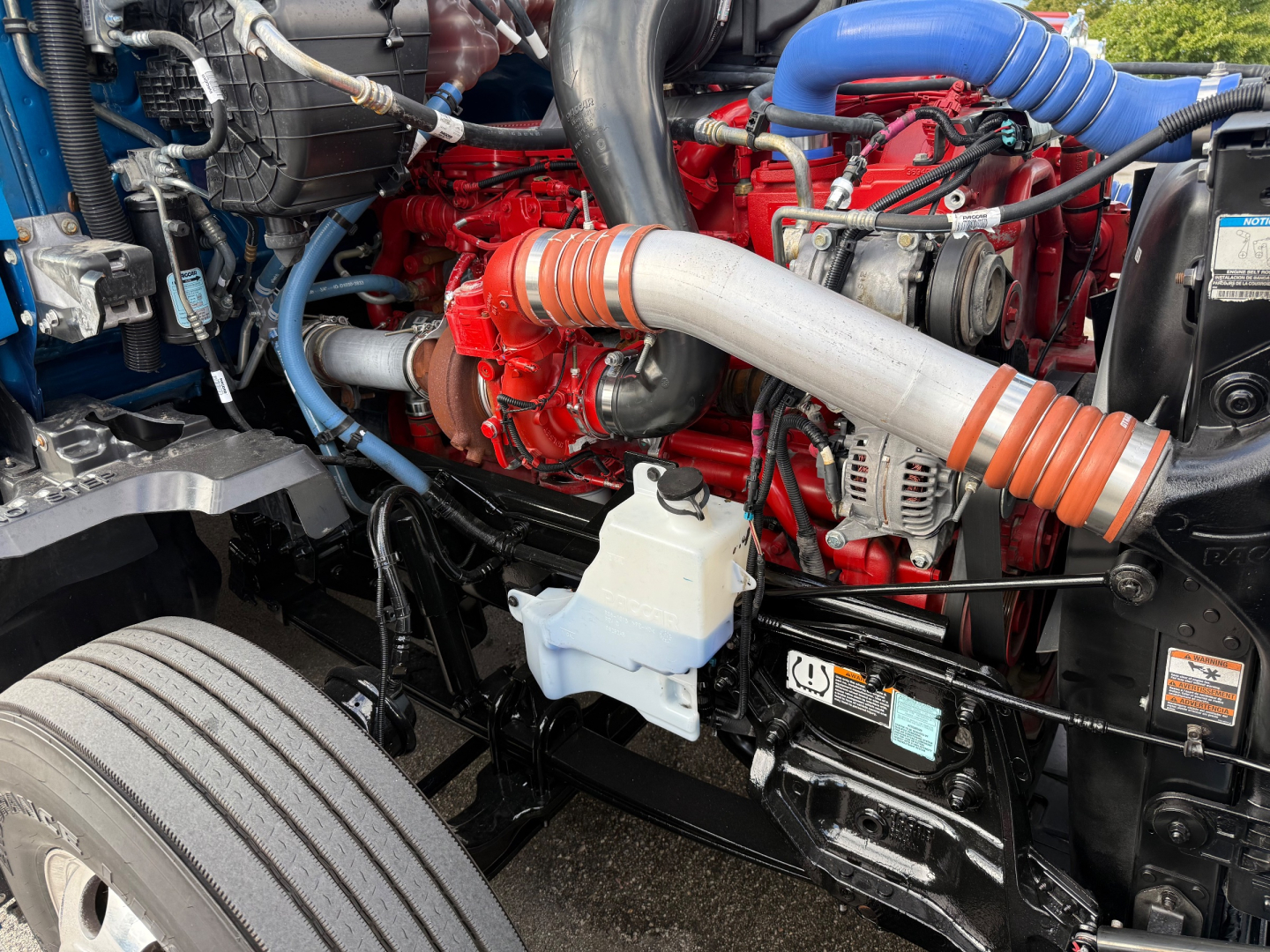 Kenworth T680 engine bay showing red engine block, large intake turbo piping, blue coolant hose, white reservoir, and various hoses and components.